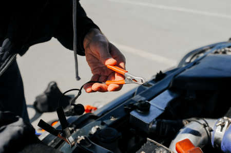 the hand connects the tongs red terminal to the battery of a car with an open hood and tuned parts...
