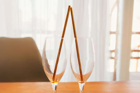 Two beautiful champagne glasses with gold paper tubes on a wooden table against the background of a large window with light curtains. Romantic date for two at homeの写真素材