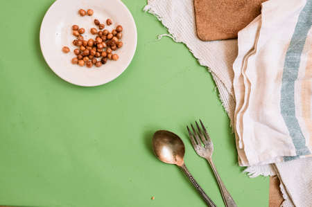 white plate with ready-baked chickpeas on a cotton vintage napkin and an old metal fork and spoon on a clean green background with scattered grains of raw chickpeas. Content for healthy eating.の写真素材