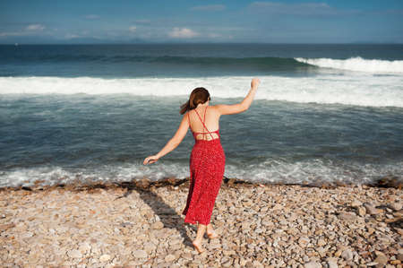 young slender girl in a red long dress with an open back walks by the ocean with large waves on her...