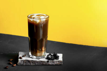 cold black coffee in high glass on stand of natural stone, coffee beans and ice cubes on black table and yellow backgroundの写真素材
