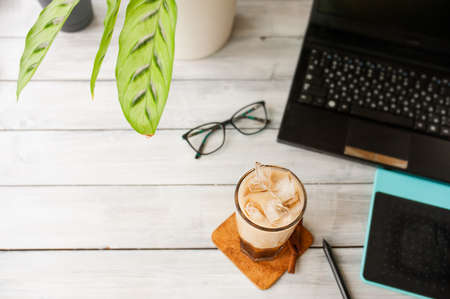 cold coffee with ice and milk on working office light table with computer, graphic tablet, glasses, calathea plant, top view, space for textの写真素材