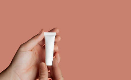 hands with delicate skin and neat nude manicure hold small white clean cosmetic tube on delicate pink background, isolated skin care layoutの写真素材