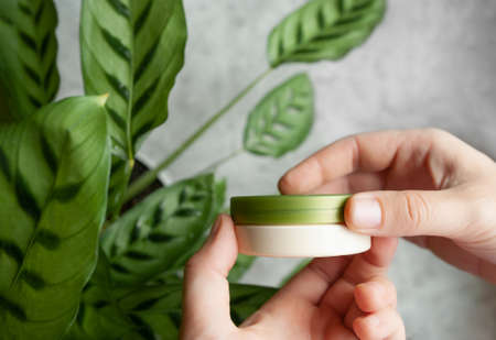 hands with neat manicure unscrew the green lid from beige jar of cream with green plant on...