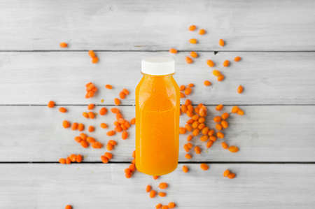 bottle of bright orange detox drink with white closed lid against the background of scattered sea buckthorn berries on a light wooden tableの写真素材