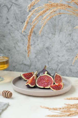 fresh ripe slices of figs on stylish beige ceramic plate, bowl with honey and honey stick, gray napkin and bouquet of dried flowers. cozy healthy breakfast, vertical, selective focusの写真素材