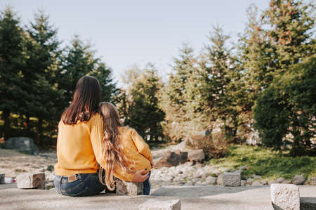 mother and daughter with long blonde hair in identical yellow knitted warm sweaters tenderly embrace and admire autumn forest and trees in stone garden, rear viewの写真素材