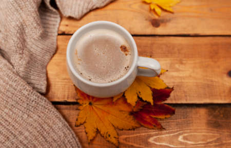coffee with milk foam in a white mug, autumn red and yellow maple leaves and a knitted beige sweater on a wooden table, top view, monochrome cozy picture of Breakfastの写真素材