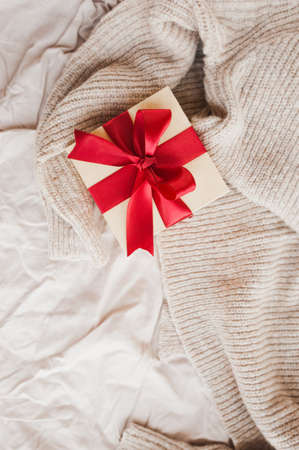 stylish square beige box with red festive satin ribbon on beige cozy crumpled blanket selective focusの写真素材