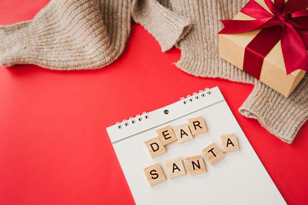 white spiral notebook and square inscription dear Santa on red background, gift box made of Kraft paper with red satin bow, garlands and knitted fabric on background, selective focusの写真素材