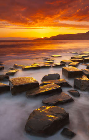The sun sets on the beautiful Dorset coastline illuminating glistening rocks with orange and yellow highlightsの写真素材