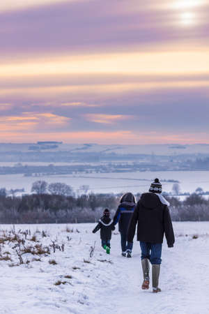As sunset approaches the mist hangs in valleys over snowy, Oxfordshire fields near Wittenham Clumps. A family walk through the snow towards the sunset.の写真素材
