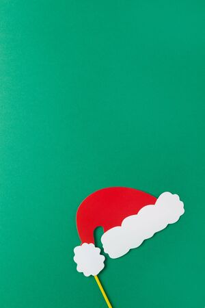 Christmas decoration, red Santas hat on stick on green background with copy space. Festive concept, party accessories. Vertical, flat lay. Minimal style. Top view.の写真素材