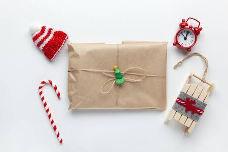 Christmas gift wrapped in brown craft paper, tied with scourge, with cane candy, small analog clock, sled, hat on white background. Minimal style. Top view. Do it yourself, celebration, eco conceptの写真素材
