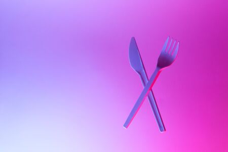 Disposable appliances plastic white fork and knife levitate in neon light, pink blue background, copy space. Eco, zero waste concept. Front view. Horizontal.の写真素材