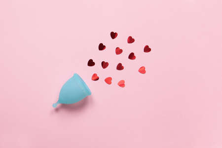 Turquoise menstrual cup on pastel pink background with red hearts confetti. Concept zero waste,...