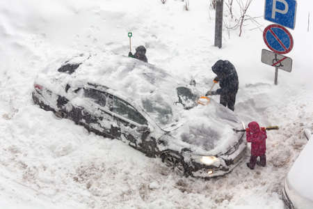 Unrecognizable people are clearing snow-covered car into snowstorm. Children help their father to clear snow from car. There is strong blizzard outside. Winter routine and problems concept.の写真素材