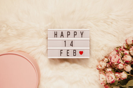 Light box with inscription Happy 14 FEBRUARY, bouquet of small roses, gift box on white fur carpet. Valentine's Day. Flat lay. Top view. February 14th celebration concept. Minimal style. Toned.の写真素材