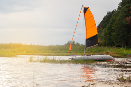 Inflatable sailing catamaran with raised sails on the river near the shore. Summer sports concept, fishing, outdoor recreation, activity, travel, vacation. Side view, sun flare. Horizontal.の写真素材