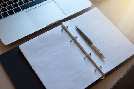 Top view on working place. Laptop and open notebook with pen on beige table. Notepad sheets on silver brackets, automatic ballpoint pen in silver-black color. Business concept. Soft selective focus.の写真素材