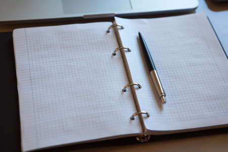 Top view on working place. Laptop and open notebook with pen on beige table. Notepad sheets on silver brackets, automatic ballpoint pen in silver-black color. Business concept. Soft selective focus.の写真素材