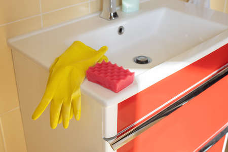 Cleaning bathroom sink and faucet with detergent in yellow rubber gloves and pink sponge. Concept housework, housekeeping, hygiene, service of cleaning. Light cleaning methods. Copy space. Side view.の写真素材