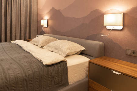 Modern bedroom in the apartment, in neutral earthy tones. Double bed with a soft textile headboard. Beautiful bedside lamps shine with warm light. Minimalism style, Scandinavian. Horizontal.の写真素材