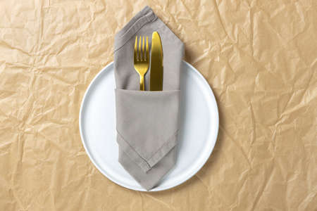 Cutlery, gold fork and knife in folded cloth napkin on white round plate on crumpled craft paper, minimal style flat lay, copy space. Table setting concept, environmentally friendly food, zero waste.の写真素材