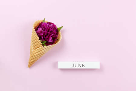 Peony flower in waffle cone, wooden block with inscription June on pastel lilac background, copy space. Minimal style flat lay. For greeting card, invitation, birthday. Summer concept.の写真素材