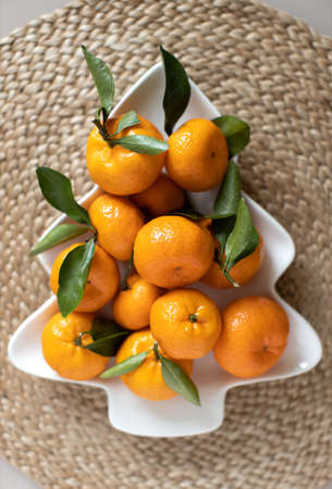 Small ripe tangerines with leaves, on plate in shape of Christmas tree on wicker serving napkin. Concept winter vitamin food. Moody still life festive composition. For greeting card, book, stories.の写真素材