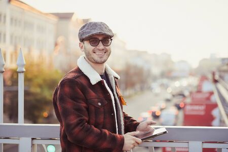 Young man in dark glasses grey cap brown jacket standing on city bridge over highway street smiling...