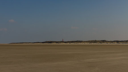 A lone red lighthouse stands against rolling sand dunes and an open blue sky, offering a striking contrast to the vast, empty beach, creating a sense of solitude and remoteness. Captured in Schiermonnikoog National Park, Netherlands, Europe.の写真素材