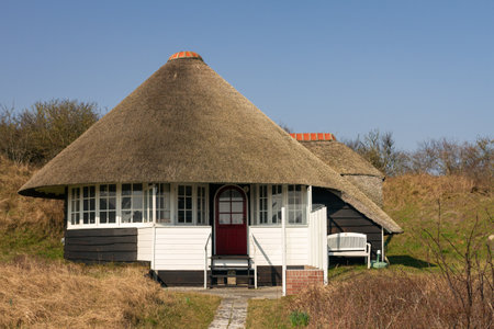 A charming thatched-roof cottage with white accents and a red door sits amidst rolling dunes, blending harmoniously with the surrounding landscape under a clear blue sky. Captured in Schiermonnikoog National Park, Netherlands, Europe.の写真素材