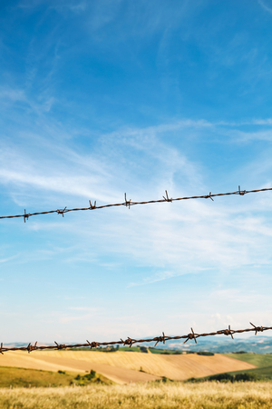 Fence with barbed wire, fields and blue sky on backgroundの写真素材