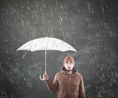 Young man under an umbrella with rain drawn on a blackboard in the background over himの写真素材