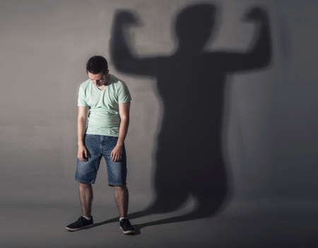 Image of a weak man standing depressed in front of a wall and his shadow shows streghtの写真素材