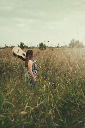 Young girl holding guitar on her back and walking in the field.の写真素材