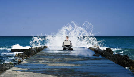 Young man working on laptop sitting on a breakwater , big waves breaking behind him.の写真素材