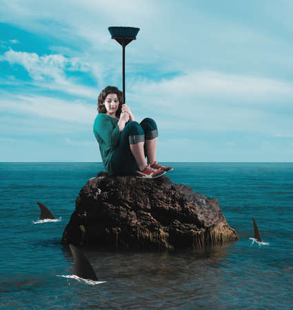 Scared woman protect herself from sharks with a broom on a rock in the ocean.