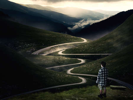 Man standing on a mountain peak admiring the view of a curved road crossing the mountains.
