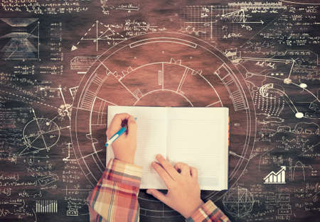 Top view of hands writing on a notebook surrounded by math formulas and sketches drawn on wooden table.の写真素材