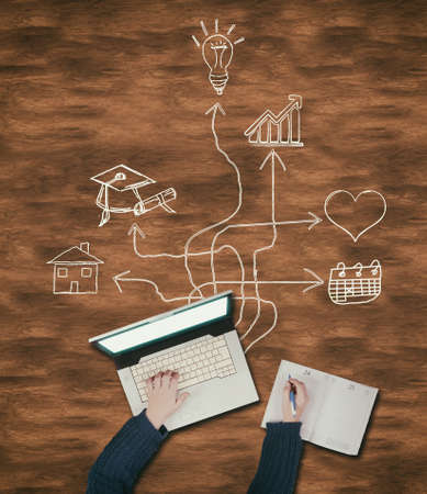 Top view of hands writing on laptop and notebook on a wooden table, drawn with arrows leading to different icons.の写真素材