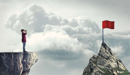 Man on a mountain peak looking through binoculars to a red flag. The concept of reaching goals.の写真素材