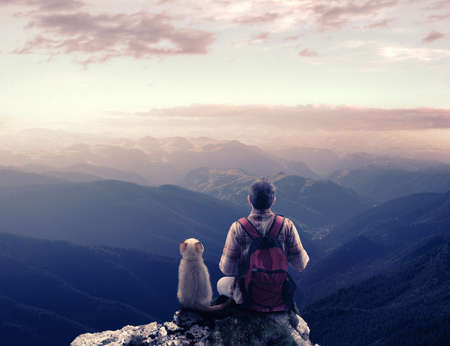 Hiker and a cat sits on top of a mountain peak admiring the view.