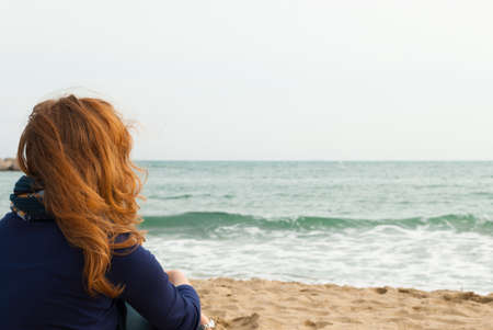 Redhead girl on a Barcelona sand beach looking at the sea, view from behindの写真素材