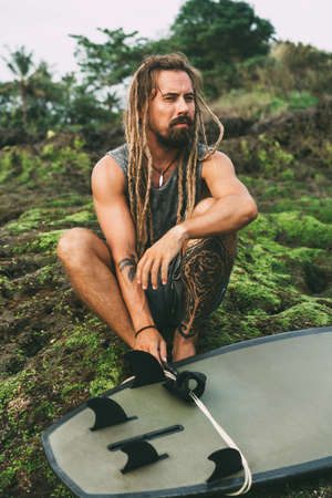 One young hipster man with dreadlocks and tattoos sitting near the ocean on the mossy rocks in...