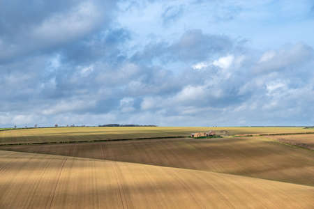 View of a deralict farm over rolling ploughed striped fields  on a sunny day with cloudy sky.の写真素材