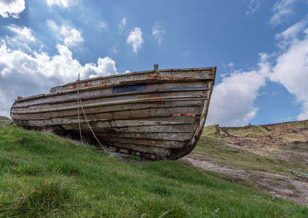 A distresed wooden fishing boat beached on grass enbankment on sunny day with blue sky background.の写真素材