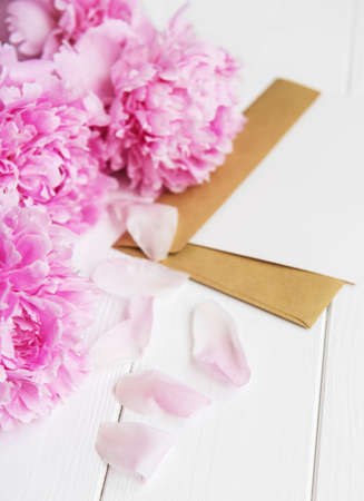 invitation card, craft envelope and pink peony flowers on a white wooden tableの写真素材