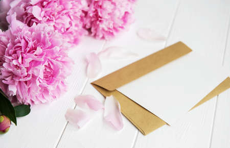 invitation card, craft envelope and pink peony flowers on a white wooden table